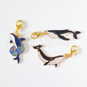 May include: Three enamel charms with gold-colored clasps. One charm is a blue and white shark with a starry design. The other two charms are black and white whales with gold-colored clasps.