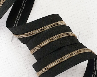 May include: Black zipper with gold-colored teeth, coiled and displayed against a white background. The zipper tape is black, and the teeth are a contrasting gold color. The zipper is likely for sewing or crafting projects.