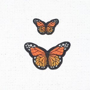 May include: Two embroidered butterfly patches, one larger than the other, with orange and black wings. The butterflies are on a white fabric background.