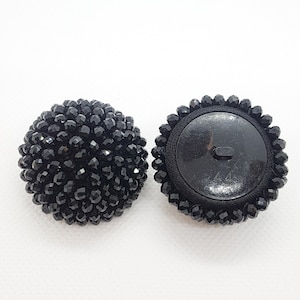 Puede incluir: Dos botones negros con cuentas. Un botón está completamente cubierto de pequeñas cuentas facetadas. El otro botón tiene una parte trasera de tela negra con un botón de plástico en el centro y un borde con cuentas. El número "44" es visible en la parte posterior.