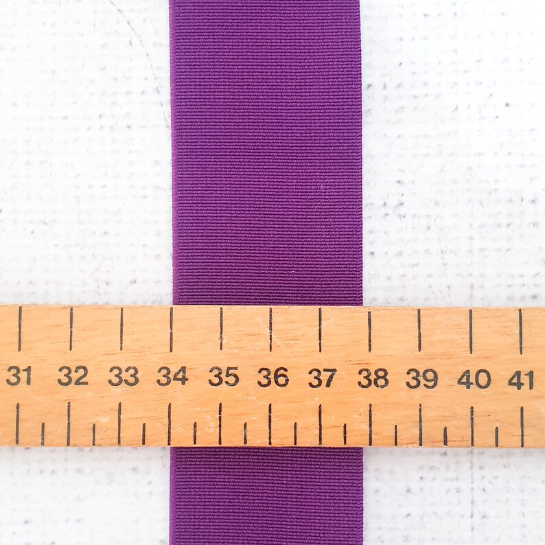 Pu&ograve; includere: Nastro elastico viola misurato contro un righello di legno con i numeri da 31 a 41. Il nastro &egrave; largo circa 2,5 cm.