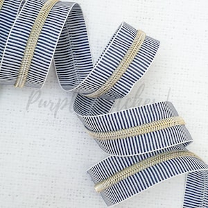 May include: A close-up of a gold zipper with a white and navy blue striped tape. The zipper is coiled and ready to be used for sewing projects.