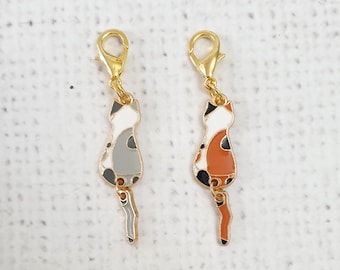 May include: Two cat-shaped charms with gold-coloured clasps. One charm is grey, white, and black, while the other is orange, white, and black. Both charms have a gold-coloured tail and are designed to be attached to a keychain or bracelet.