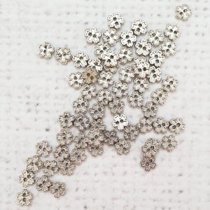 May include: A large quantity of silver metal flower-shaped embellishments. The embellishments are scattered on a white surface.