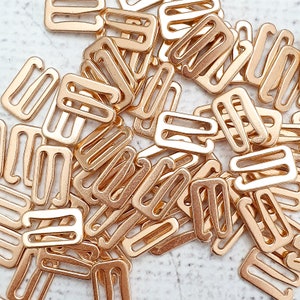 May include: A pile of gold-toned metal bra strap adjusters. The adjusters are rectangular with two loops on each side.