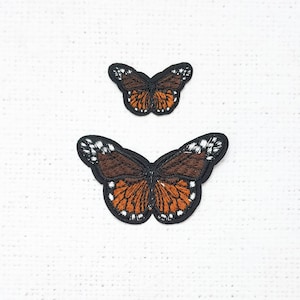 Pode incluir: Dois patches de borboleta bordados, um maior que o outro. Ambas as borboletas são pretas com asas marrons e laranja e detalhes brancos.