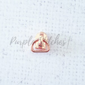 May include: Rose gold metal zipper pull with a D-ring. The zipper pull is designed to be used on a zipper for clothing or bags.