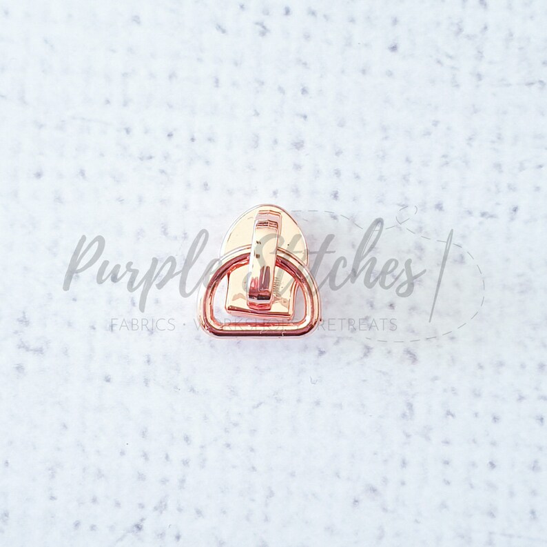 May include: Rose gold metal zipper pull with a D-ring. The zipper pull is designed to be used on a zipper for clothing or bags.