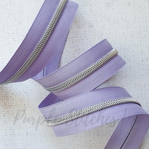 May include: A close-up of a lavender zipper with a silver metal zipper pull. The zipper is coiled and folded, showing the detail of the stitching and the metal teeth.