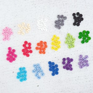 May include: Assortment of colourful two-hole buttons in various colours, including pink, red, orange, yellow, green, blue, purple, and grey. The buttons are arranged in rows on a white surface.