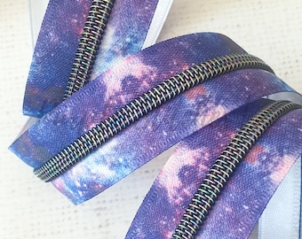 Puede incluir: Primer plano de una cremallera con un estampado de galaxia en tonos azules, morados y rosas. Los dientes de la cremallera son de color iridiscente metálico. La cinta de la cremallera está doblada sobre sí misma.