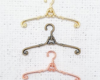 Könnte beinhalten: Drei dekorative Kleiderbügel in Gold, Bronze und Roségold. Jeder Kleiderbügel hat ein einzigartiges, verziertes Design mit einer gerollten Oberseite und einer herzförmigen Mitte.