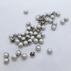 Puede incluir: Un grupo de botones blancos con forma de perla con engastes de metal plateado. Los botones están esparcidos sobre una superficie blanca.