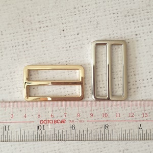 Pu&ograve; includere: Due fibbie scorrevoli in metallo, una dorata e una argentata, che misurano circa 2,5 cm di lunghezza. Le fibbie sono mostrate su un righello bianco.