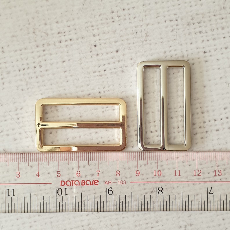 Pu&ograve; includere: Due fibbie scorrevoli in metallo, una dorata e una argentata, che misurano circa 2,5 cm di lunghezza. Le fibbie sono mostrate su un righello bianco.