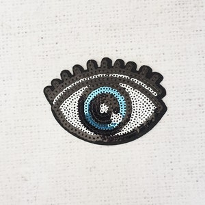 May include: A sequined eye patch with a black outline, white and gray sequins, and a blue iris. The eye patch is a decorative accessory, possibly for clothing or crafts.