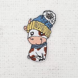 May include: Embroidered patch featuring a cartoon cow wearing a blue knit hat with a white pom-pom and a blue striped scarf. The cow has brown spots, a pink muzzle, and is sitting down.