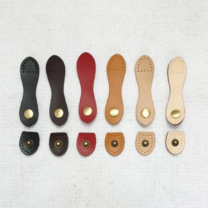 May include: Ten leather strap toggles in various colors, including black, brown, red, and beige. Each toggle has a brass button and a rounded shape.