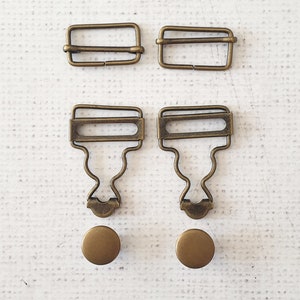 May include: A collection of antique brass-colored metal hardware components, including two rectangular buckles, two suspender clips, and two round buttons. The hardware is arranged on a white textured surface.