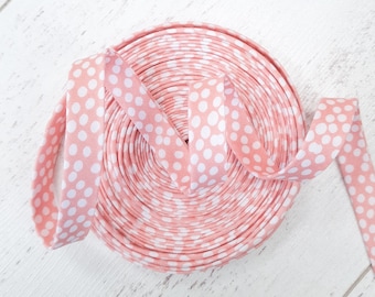 Könnte beinhalten: Eine Rolle rosa Schrägband mit weißen Polka-Dots. Das Band ist spiralförmig aufgerollt, mit einem herausragenden Stück. Der Hintergrund ist eine weiße, strukturierte Oberfläche. Geeignet für Näh- und Bastelprojekte.