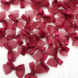 May include: A large group of burgundy-colored satin ribbon bows with a sheer overlay. The bows are arranged in a random pattern on a white surface.