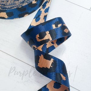 Könnte beinhalten: Ein Rolle blau-beiges Webband mit Leopardenmuster. Das Gurtband hat einen marineblauen Hintergrund mit schwarzen und beigen Leopardenflecken. Das dicht gewebte Gurtband scheint haltbar zu sein. Die Rolle ist teilweise abgerollt und zeigt das Muster.