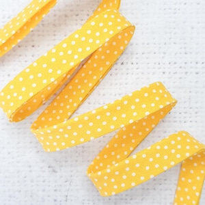 Peut inclure: Ruban jaune vif à pois blancs. Le ruban est plié et disposé en motif ondulé sur un fond blanc texturé. Ce ruban est adapté aux travaux manuels et à l'emballage de cadeaux.