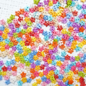 May include: A large collection of colourful star-shaped buttons in various shades of pink, blue, green, yellow, orange, and red. The buttons are scattered on a white surface.
