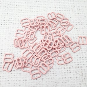 Puede incluir: Un conjunto de deslizadores de tirantes de sujetador de color rosa pálido, dispersos sobre una superficie blanca texturizada. Estos deslizadores metálicos tienen forma rectangular con dos ranuras paralelas, utilizados en lencería.