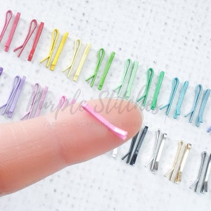 May include: A collection of colorful metal hair clips in various shades of blue, green, yellow, red, and purple. A single pink hair clip is shown on a finger.