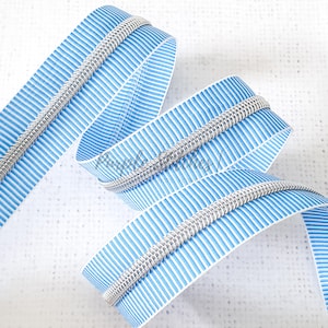 May include: Close-up of three sections of a blue and white striped zipper with a silver metal zipper track. The zipper tape is a light blue color with thin white stripes. The zipper is likely for sewing or crafting projects.