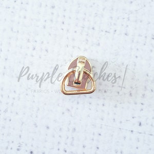 May include: Gold metal zipper pull with a pink plastic slider. The zipper pull has a D-shaped ring.