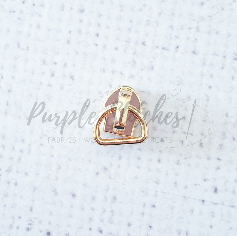 May include: Gold metal zipper pull with a pink plastic slider. The zipper pull has a D-shaped ring.