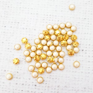 May include: A large group of gold and white pearl-like buttons. The buttons are arranged in a loose cluster on a white surface.