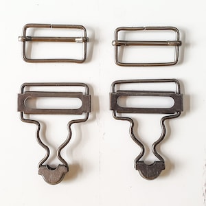 May include: Four antique metal suspender clips and buckles. Two are rectangular buckles, and two are clips with a curved design and a clasp. The metal has a tarnished, aged appearance, suggesting vintage or repurposed use.