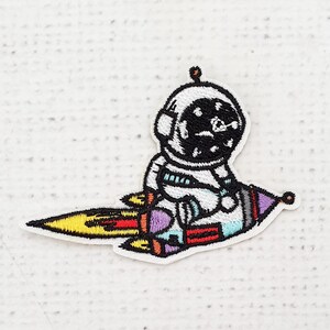 Puede incluir: Parche bordado con un astronauta de dibujos animados montado en un cohete. El astronauta lleva un traje espacial blanco y un casco negro. El cohete tiene llamas coloridas y detalles en amarillo, rojo, morado y azul.