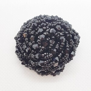 Op de afbeelding: Een ronde zwarte kralen broche met een textuur. De broche is gemaakt van kleine zwarte kralen en heeft een glinsterende afwerking.