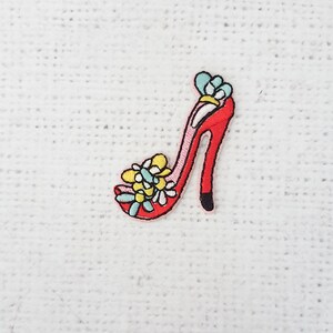 May include: Embroidered patch of a red high-heeled shoe with floral accents in yellow, blue, and white. The shoe has a black heel and a pink outline. The patch is likely for clothing or accessories.