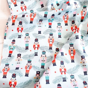 Puede incluir: Una tela azul claro con un patrón repetido de adornos de cascanueces. Los cascanueces son de varios colores, incluyendo rojo, rosa y azul. La tela está decorada con ramas de pino verdes y estrellas rojas.