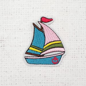 May include: Embroidered patch of a sailboat with a pink sail, blue hull, and a red anchor. The sail has yellow and green stripes. A small red flag is at the top of the mast. The patch is on a white background.