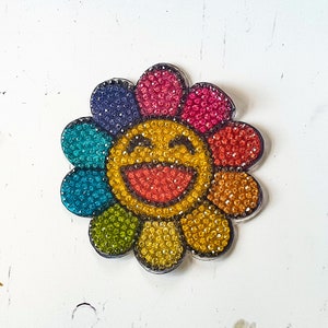 Puede incluir: Un pin de forma de flor colorido con una cara sonriente en el centro. La flor está hecha de pedrería en tonos de azul, verde, amarillo, rojo y rosa.