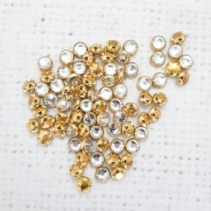 May include: A large group of clear glass rhinestones with gold settings. The rhinestones are scattered on a white surface.