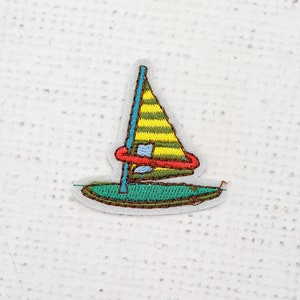 May include: Embroidered patch of a sailboat. The sail is yellow and green striped, the mast is blue, and a red ring encircles the sail. The boat's hull is green, with a white border.
