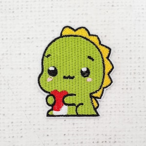 May include: Green embroidered patch of a cartoon dinosaur holding a red heart. The dinosaur has yellow spikes on its back and a cute expression.