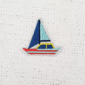 May include: A blue and white sailboat with a yellow cabin embroidered on a white fabric background.