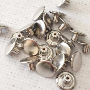 Puede incluir: Una colección de cierres a presión de metal plateado, también conocidos como poppers o botones a presión, en varios tamaños. Los cierres a presión están dispuestos en un patrón aleatorio sobre una superficie blanca.