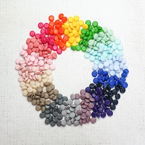 Puede incluir: Una disposición circular de cuentas planas de colores. Las cuentas están dispuestas en un espectro de arcoíris, que va del rosa al morado, pasando por el rojo, el naranja, el amarillo, el verde y el azul. Sobre un fondo blanco texturizado.