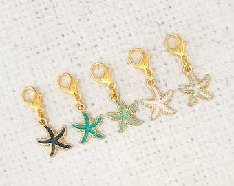 May include: Five gold-colored lobster clasp charms, each with a starfish pendant in different colors: black, teal, mint green, pink, and light blue. The starfish charms have a gold outline and are displayed on a white background.