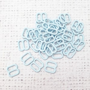 Puede incluir: Un conjunto de deslizadores metálicos de color azul claro, cada uno con forma rectangular y tres aberturas. Los deslizadores están dispersos sobre una superficie blanca texturizada. Estos pequeños componentes se utilizan probablemente en proyectos de costura o artesanía.
