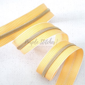 May include: A close-up of a yellow and white striped zipper with a gold metal zipper pull. The zipper is coiled and ready to be used in a sewing project.